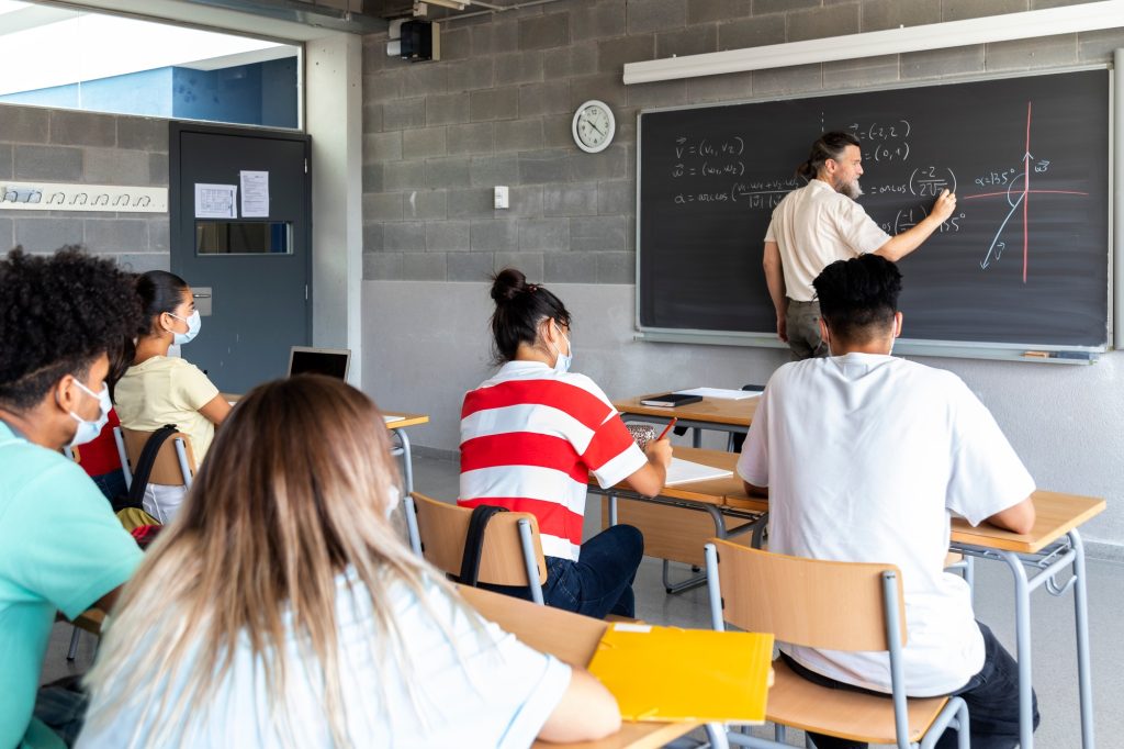 Teacher writes math formulas on blackboard. Multiracial group of students pay attention.
